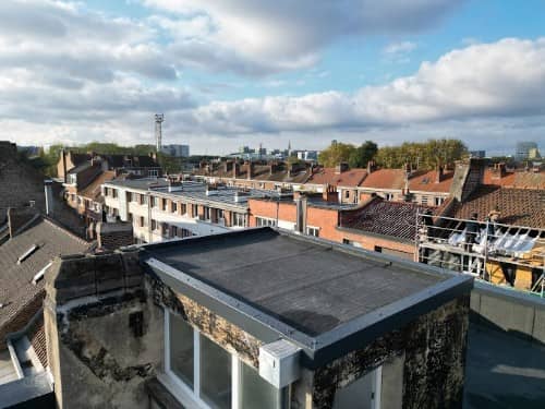 Petit toit plat d'extension recouvert d'une membrane bitumineuse et bordé de rives en aluminium, au milieu des toits d'un quartier de maisons