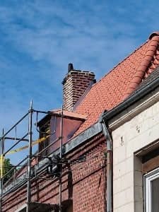 Toiture en tuiles rouges avec chien-assis et cheminée en brique rénovés, échafaudage dressé en façade